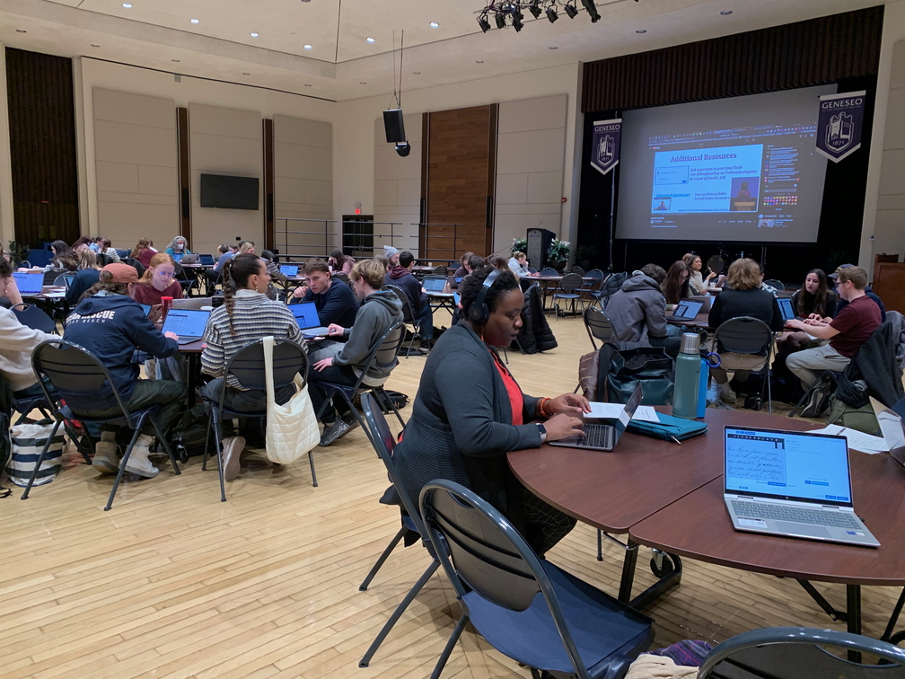 Transcribers in the College Union Ballroom seated at tables with screen showing live stream